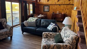 A cozy cabin interior with a wooden interior, a sofa, a patterned armchair, a dining table, and a staircase. - Riverfront 2-bedroom house tons of light, AC, deck & patio Sleeps 8 Pet Friendly (Fort Edward)
