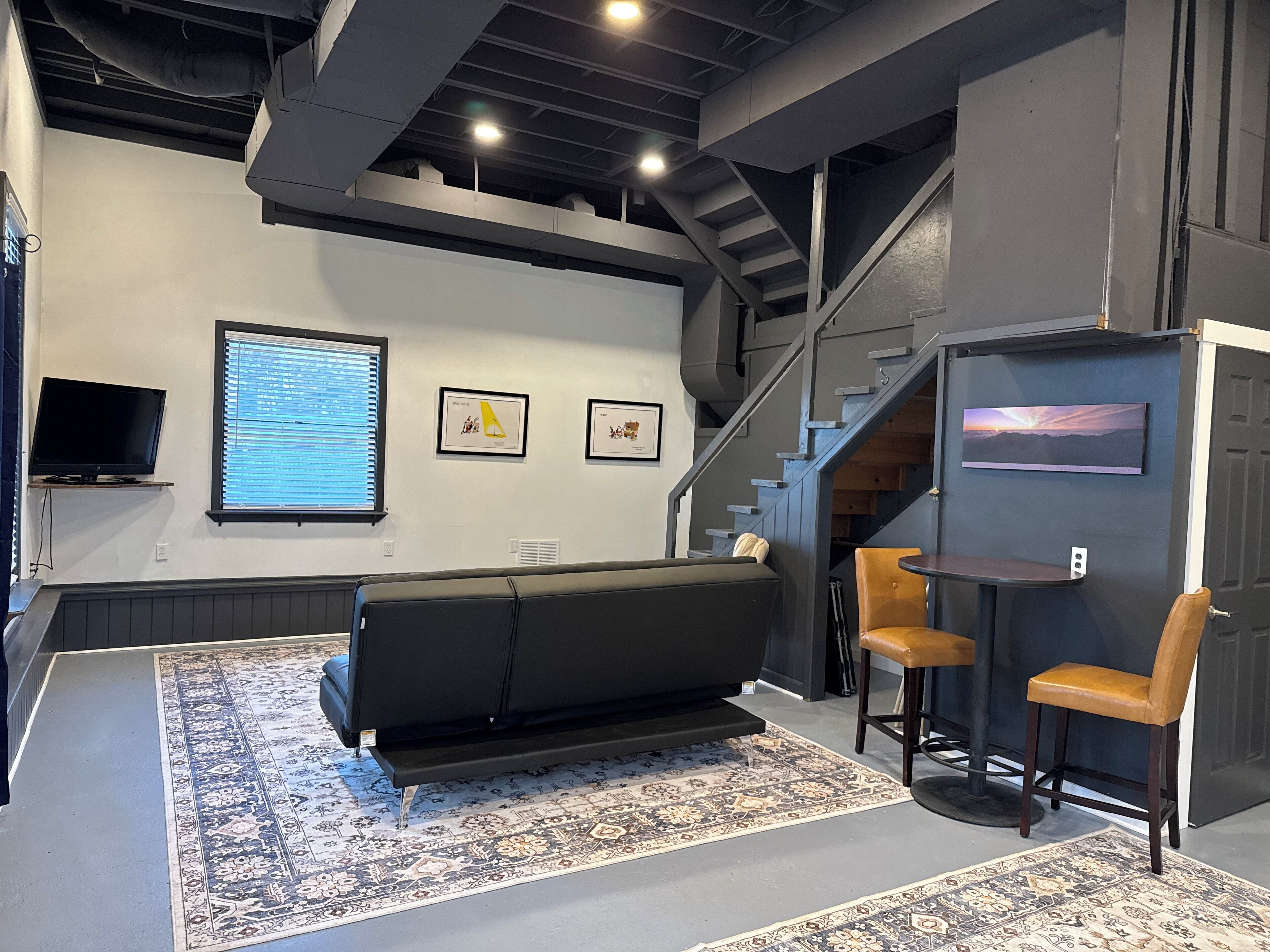 Un espace de détente moderne comprenant un canapé, deux tabourets de bar, un téléviseur à écran plat, des œuvres d’art encadrées et un escalier.
