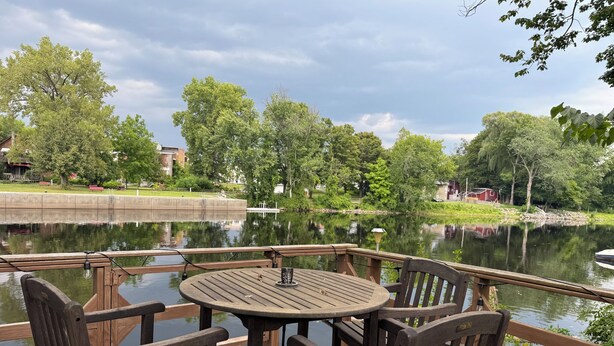 A wooden deck with a round table and chairs overlooking a calm lake, surrounded by lush greenery and trees. - Riverfront 2-bedroom house tons of light, AC, deck & patio Sleeps 8 Pet Friendly (Fort Edward)