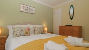 Un lit bien fait, recouvert d’une courtepointe jaune, agrémenté de deux coussins à motifs floraux et d’une couverture blanche pliée au pied du lit. On aperçoit une commode en bois avec un miroir, et un tableau encadré est accroché au mur.
