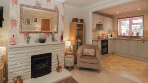 Un salon avec une cheminée, un mur miroir, un papier peint à motifs floraux, un fauteuil confortable et une cuisine avec une fenêtre.