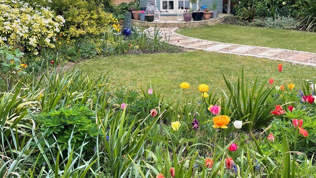 Un jardin où s’épanouissent une grande variété de fleurs et de plantes, un sentier menant à une maison à l’extérieur bleue et aux portes blanches, et une clôture en bois en arrière-plan.