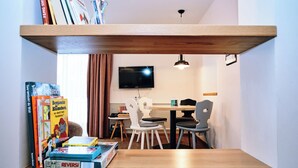 Een knus interieur met een houten plank vol boeken, een tafel met stoelen, een aan de muur bevestigde televisie en een raam met gordijnen.