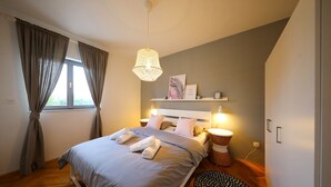 A bedroom with a bed, bedside lamps, a bedside table, a window with curtains, and a wall-mounted shelf.