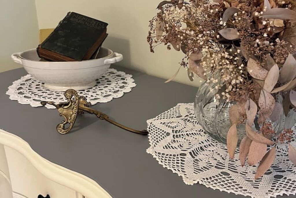 Una mesa con un cuenco blanco que contiene un libro, un mantel de encaje, un florero con flores secas y un objeto metálico decorativo.