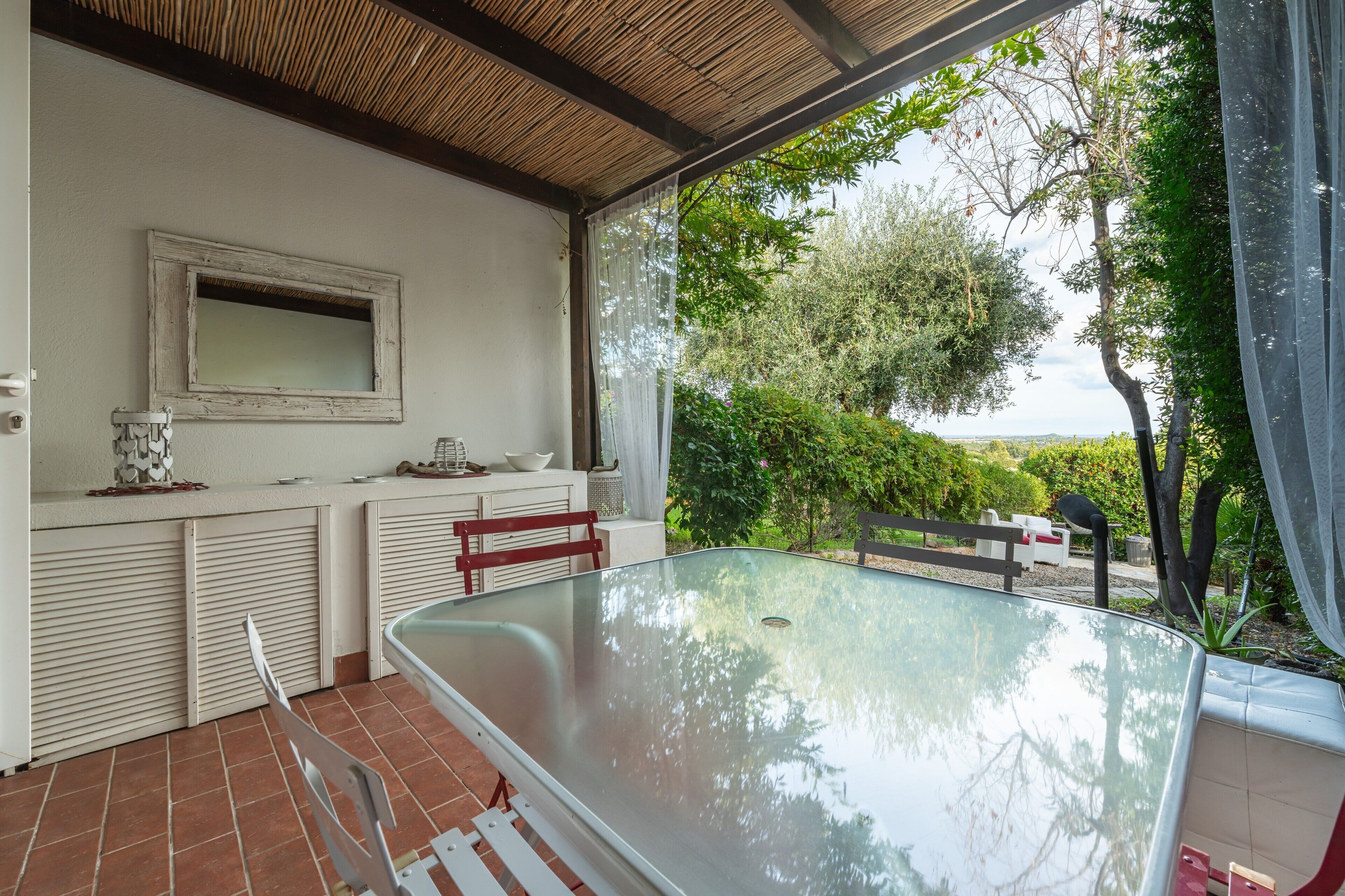 Un espace de restauration en plein air avec une table en verre, des chaises, un banc blanc, une petite table avec un saladier, un miroir et une vue sur la verdure et les arbres.