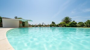 Una piscina cristalina con un muro de piedra y una zona cubierta, rodeada de exuberante follaje y palmeras.