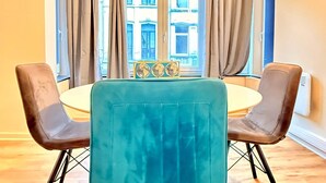 En matplats med ett runt bord, fyra stolar, en turkosklädd fåtölj och utsikt över byggnader genom ett fönster med gardiner.