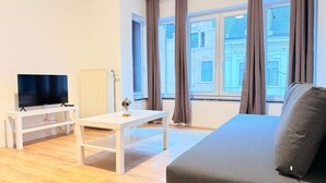 A modern living room with a flat-screen TV, a white table, a gray sofa, and large windows with curtains. - Orléans 41 (C) city center-train station-airport (Charleroi)