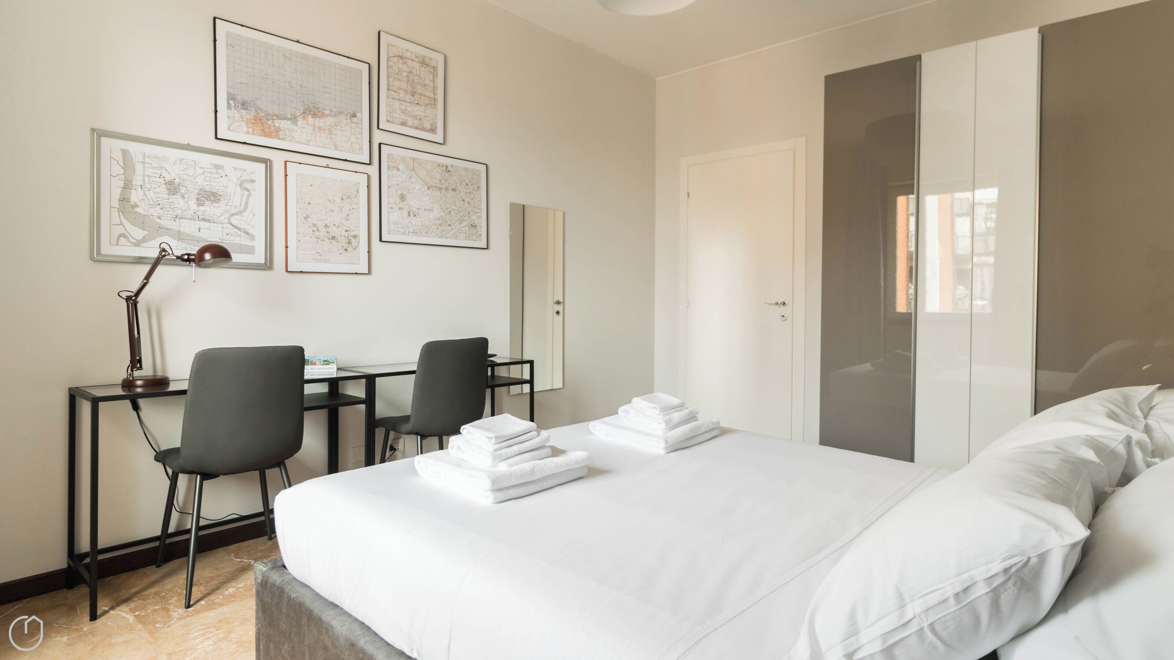 A hotel room with a bed, two chairs, a desk, a lamp, and four framed maps on the wall.