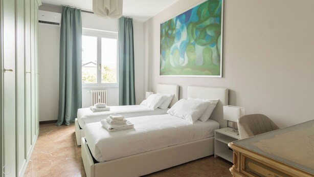 Une chambre d’hôtel avec deux lits, un radiateur, une fenêtre avec des rideaux, un tableau au mur et un bureau avec une lampe.