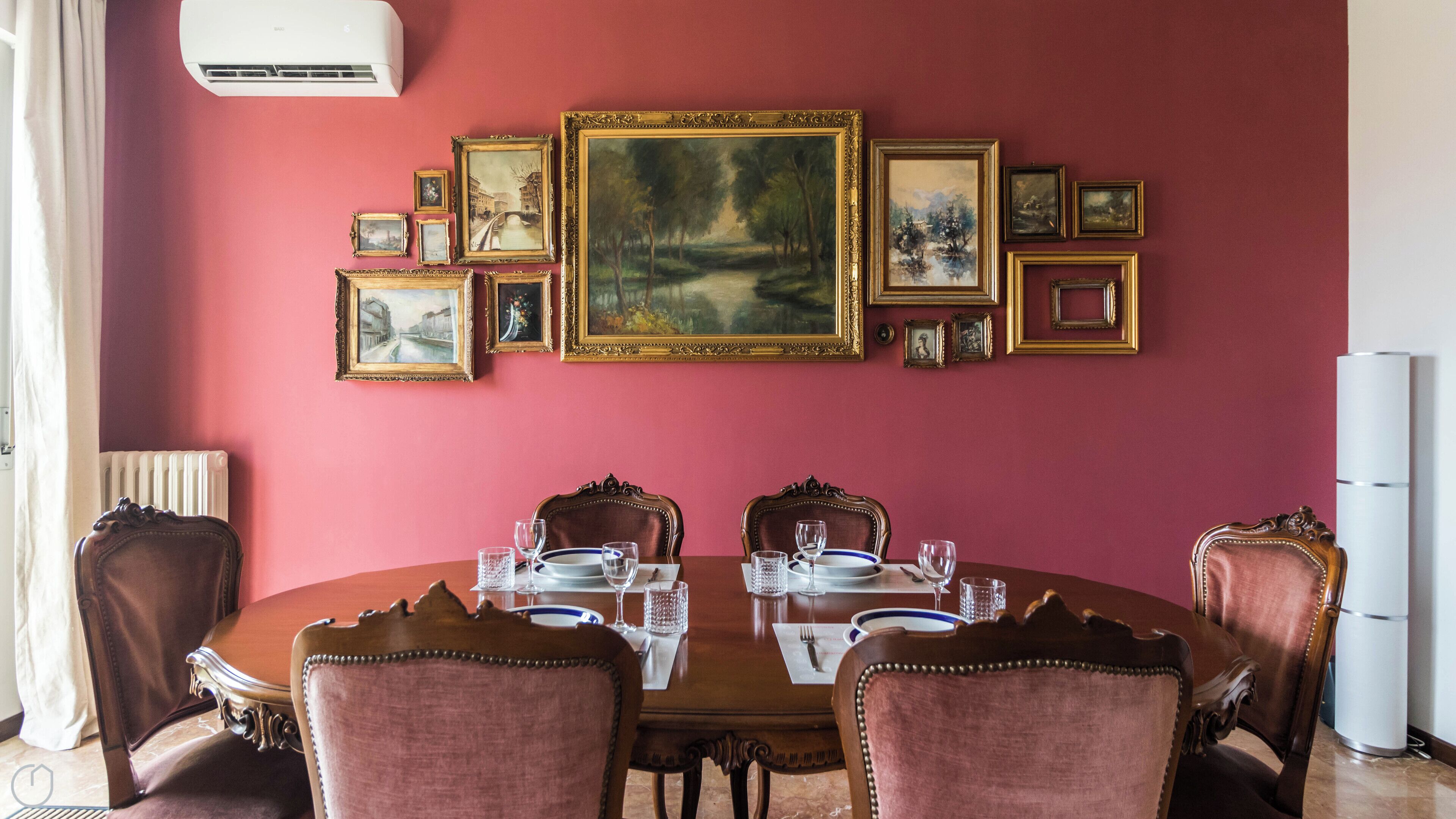 A dining room with a round table set for four, surrounded by ornate chairs, against a red wall with multiple framed artworks.