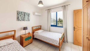 Ein Schlafzimmer mit zwei Einzelbetten, einem Holzschrank, einem Nachttisch mit Lampe, einer Deckenleuchte, einem Fenster mit Vorhängen und einer Klimaanlage.