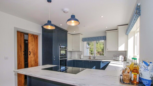 A modern kitchen with dark blue cabinets, a marble countertop, and a central island. There are two pendant lights, a window with a blue valance, and a wooden stool. - Hoddell Pitch Cottage (Presteigne)