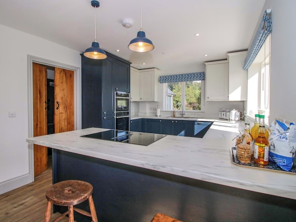 A modern kitchen with dark blue cabinets, a marble countertop, and a central island. There are two pendant lights, a window with a blue valance, and a wooden stool. - Hoddell Pitch Cottage (Presteigne)