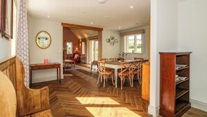 A spacious dining area with a herringbone wooden floor, a central round table surrounded by chairs, and a wooden bookcase against the wall. - Hoddell Pitch Cottage (Presteigne)