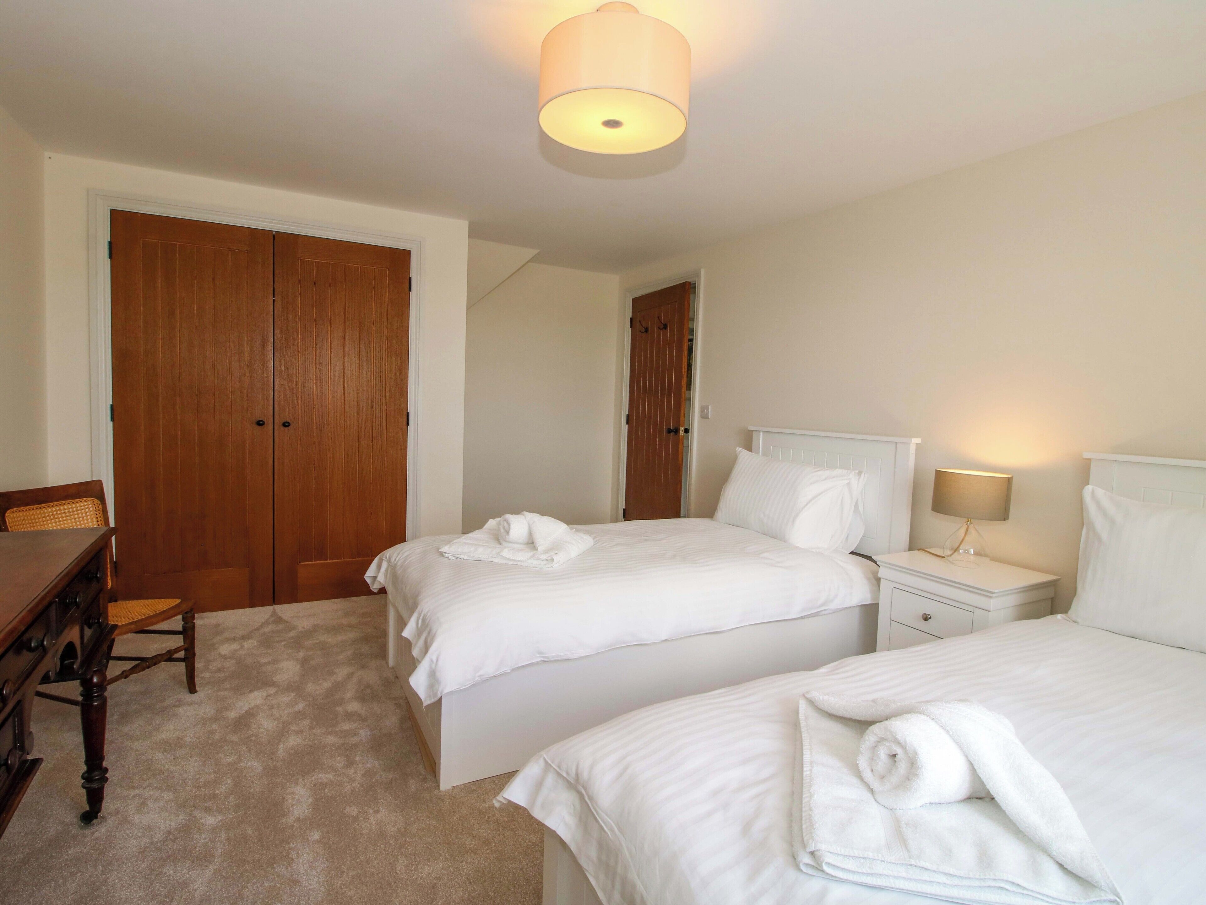 A hotel room with two beds, a wooden wardrobe, a desk with a chair, and a bedside table with a lamp.