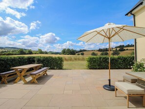 An outdoor patio with a wooden table and benches, a large umbrella, and a view of a countryside landscape. - Hoddell Pitch Cottage (Presteigne)