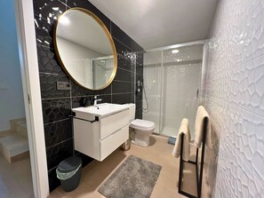 Un baño moderno con tocador blanco, un espejo redondo, un inodoro y una cabina de ducha.
