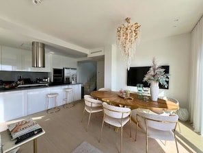 Une cuisine moderne avec des meubles blancs, une hotte en acier inoxydable et un coin repas comprenant une table en bois et des chaises dorées.