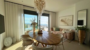 Una sala moderna con una mesa redonda de madera, sillas blancas tapizadas, una lámpara de araña, un sofá y un ventanal con vista a la ciudad.
