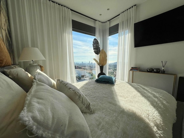 Une chambre avec un grand lit, des rideaux blancs, une vue sur le paysage urbain, une lampe et un vase contenant des plantes séchées.