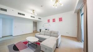 Een ruime woonkamer met een hoekbank, een ronde salontafel en een kamerbreed tapijt. Er zijn ventilatieopeningen in het plafond en een kunstwerk aan de muur bevestigd.