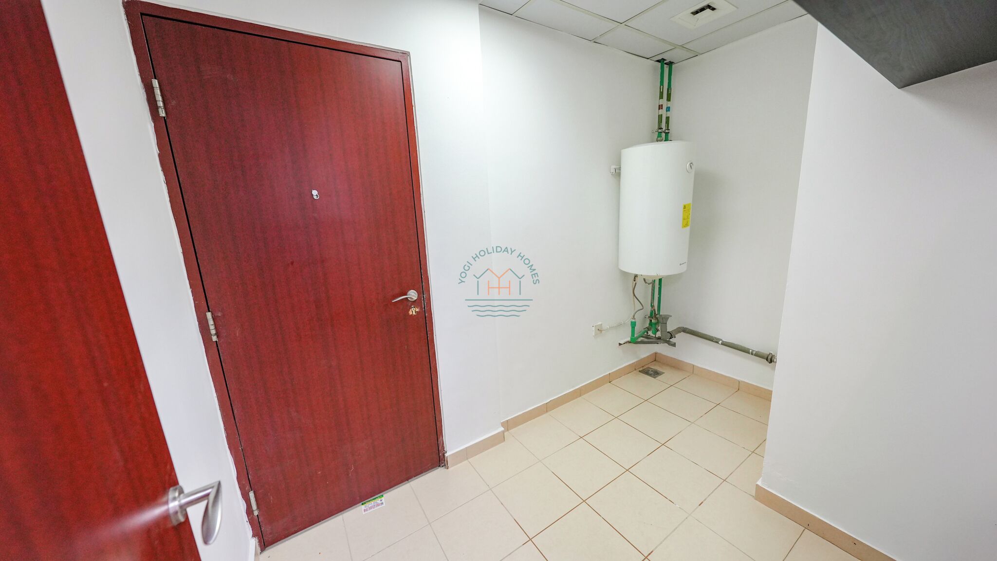 Un pasillo con una puerta roja, paredes blancas y suelo de baldosas. Un calentador de agua está montado en la pared.