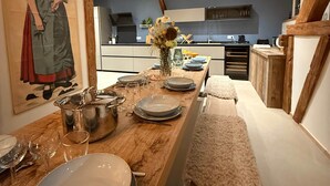 A dining area with a wooden table set for a meal, a painting on the wall, and a view into the kitchen. - La Grange - at the Ancienne ferme (Steinseltz)