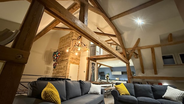 A spacious living area with a high ceiling and exposed wooden beams, featuring two grey sofas with yellow and white cushions, a glass coffee table, and a decorative hanging light fixture. - La Grange - at the Ancienne ferme (Steinseltz)