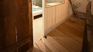 A wooden loft bedroom with a sloped ceiling, a bed with a green blanket, a bedside table with a lamp, and a window with a view of the outside. - La Grange - at the Ancienne ferme (Steinseltz)