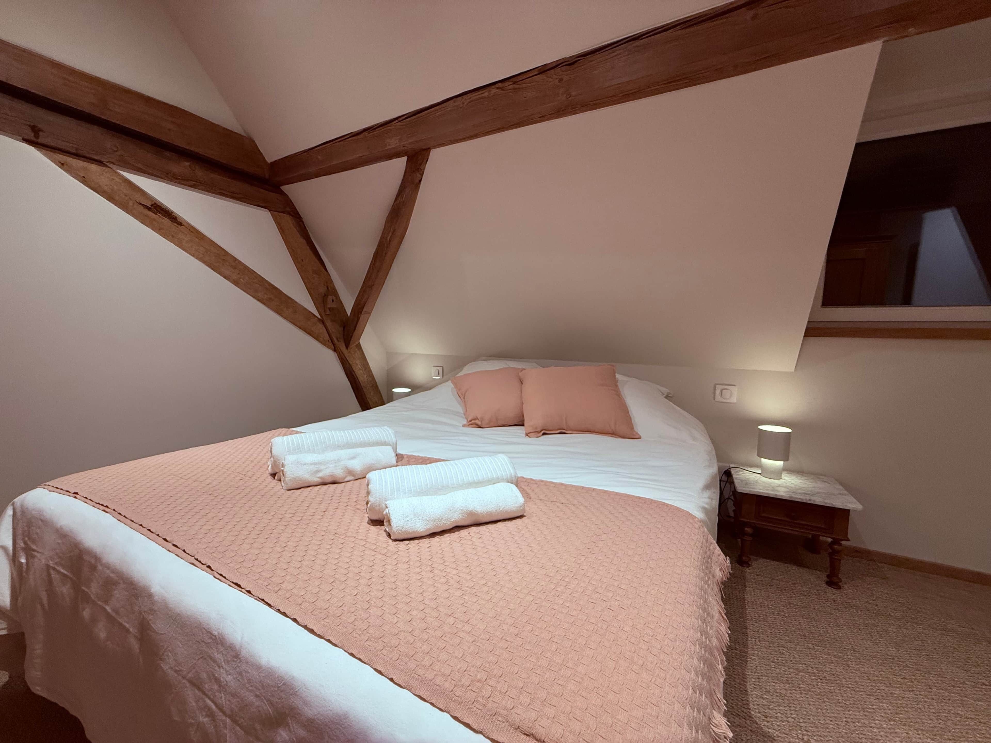 A bedroom with a high ceiling, exposed wooden beams, a large bed with white and pink bedding, two bedside lamps, and a window.