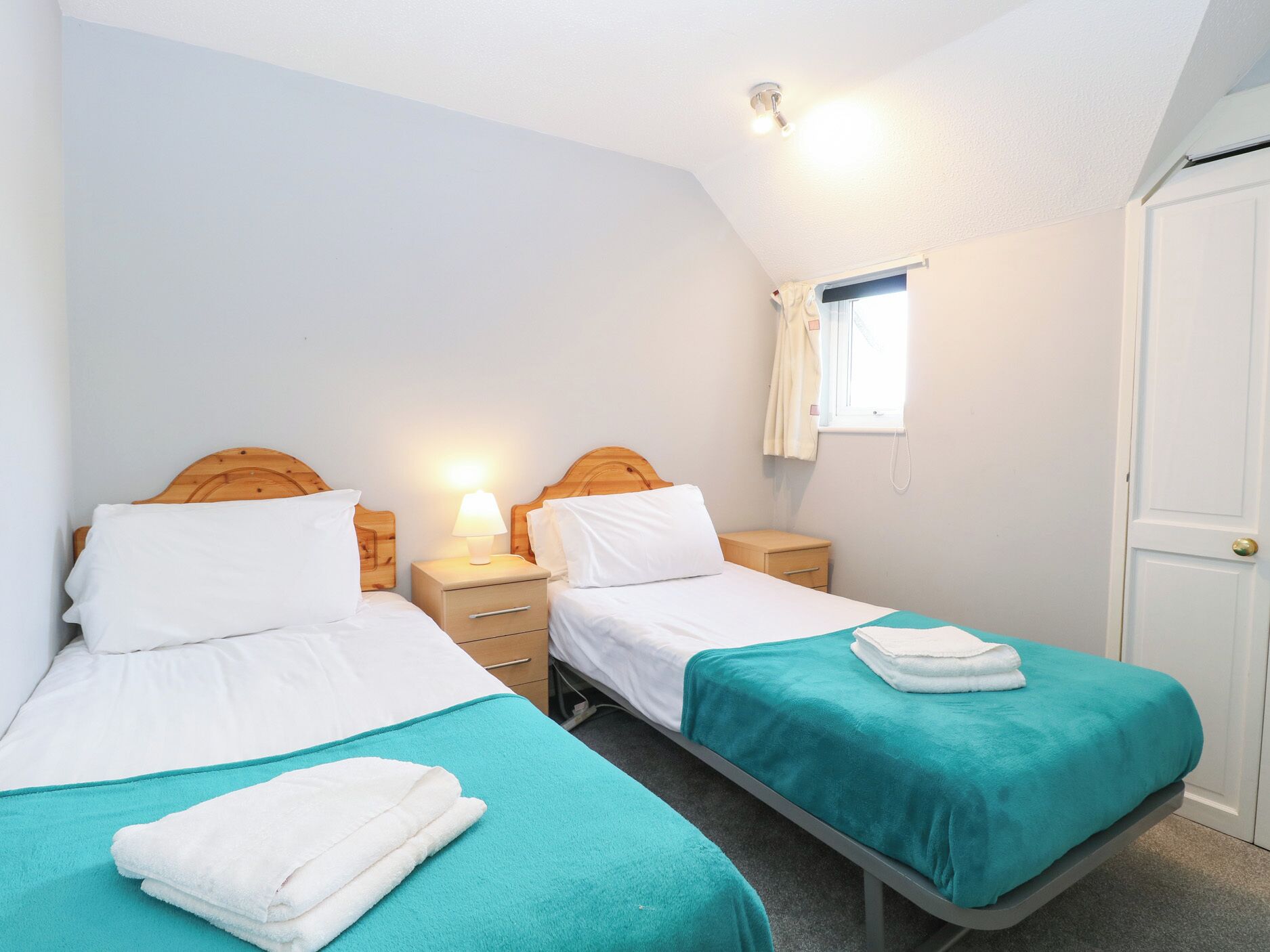Ein Doppelzimmer mit zwei Einzelbetten, jeweils mit einem Holz-Kopfteil, einem Nachttisch mit Lampe und einem Vorhangfenster.