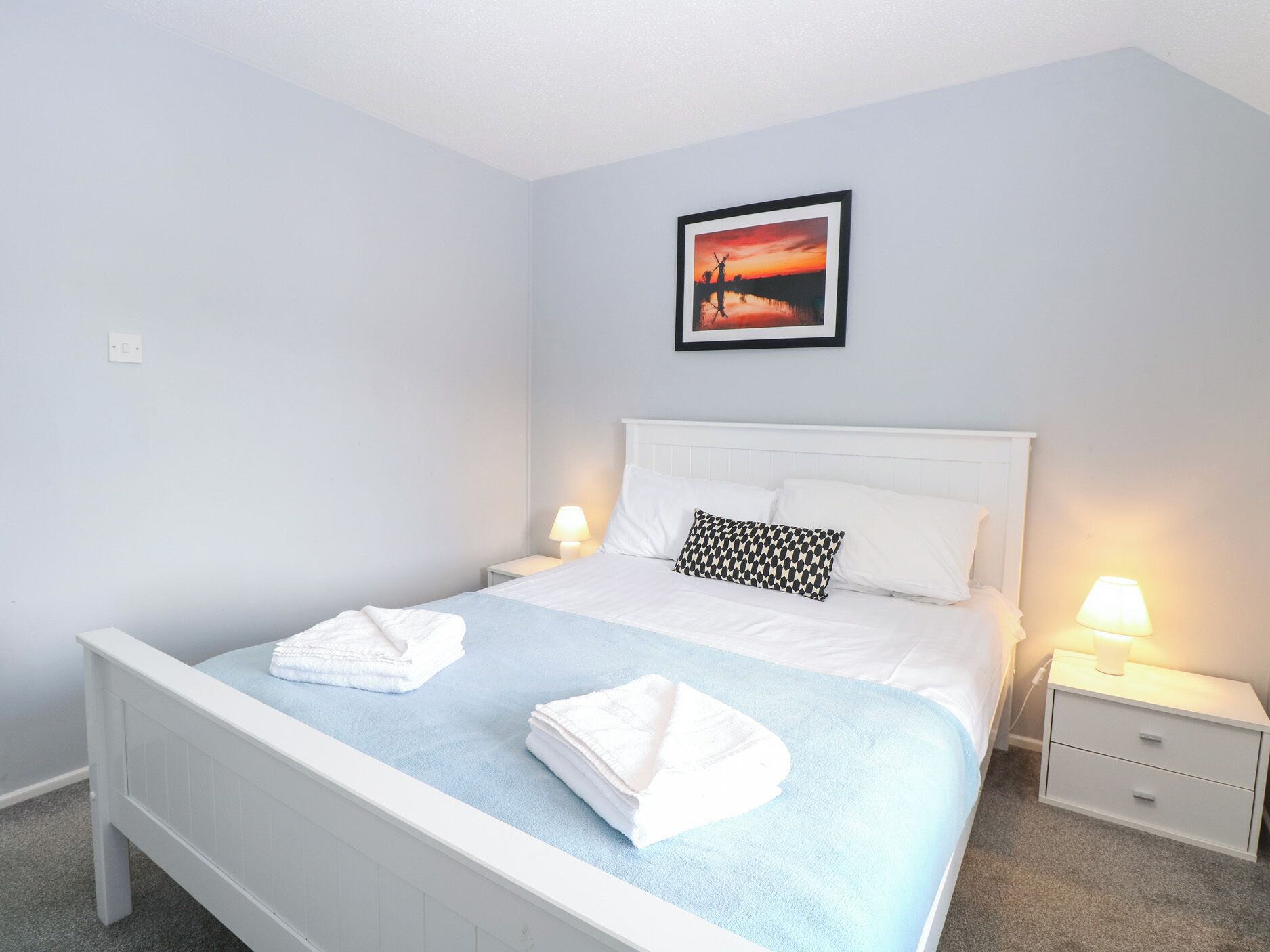 Un lit bien fait, avec du linge de lit blanc et un élément décoratif bleu, une table de chevet avec une lampe et un tableau encadré au mur.