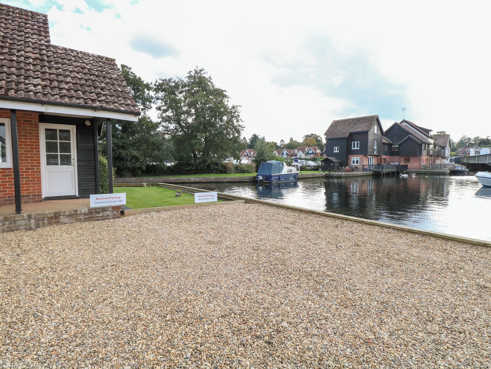 A riverside property with a gravel driveway, a boat dock, and a row of houses in the background.