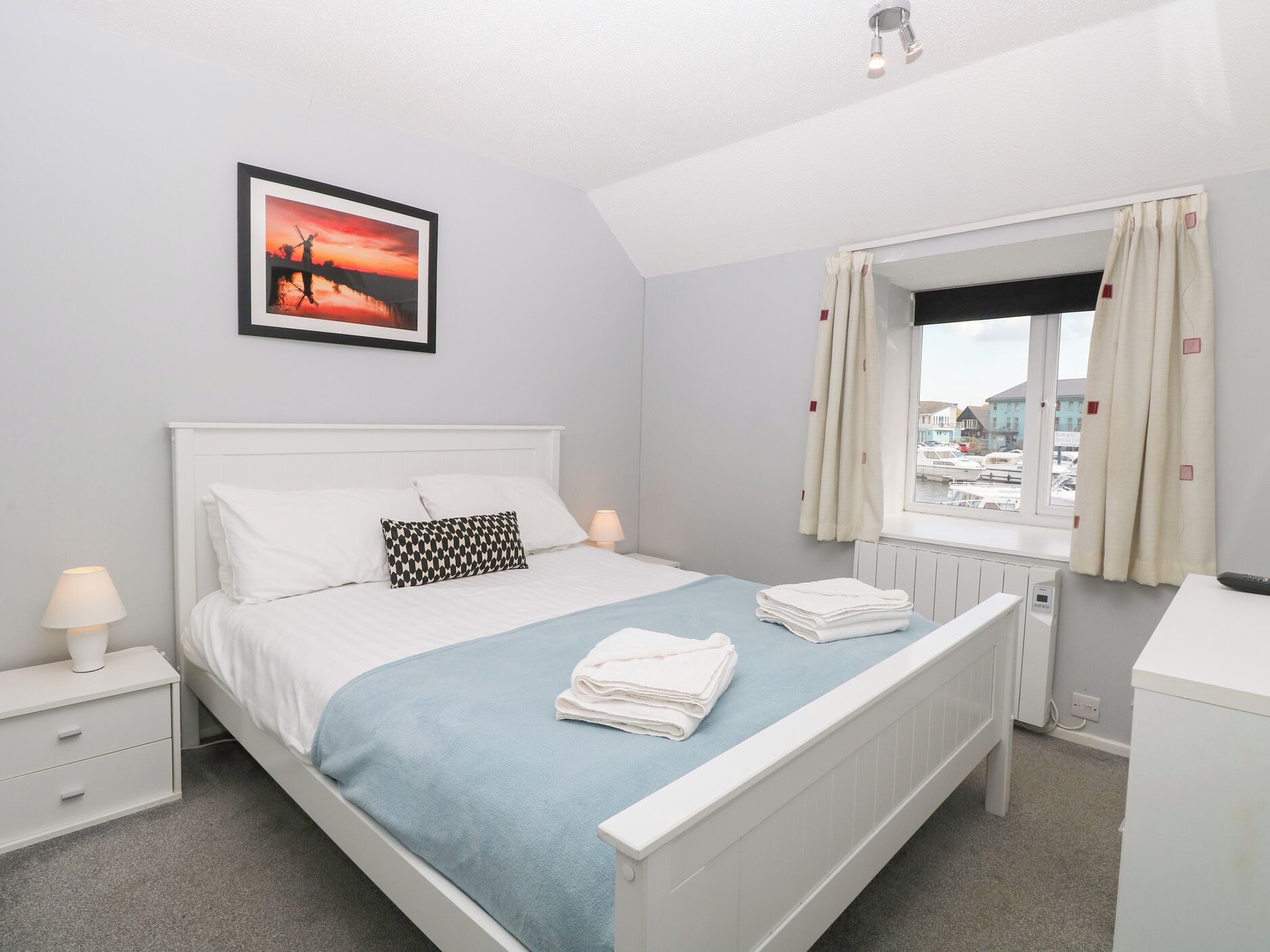 A neatly arranged bedroom with a bed, a bedside table with a lamp, a window with curtains, and a framed picture on the wall.