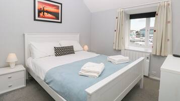 A neatly arranged bedroom with a bed, a bedside table with a lamp, a window with curtains, and a framed picture on the wall.