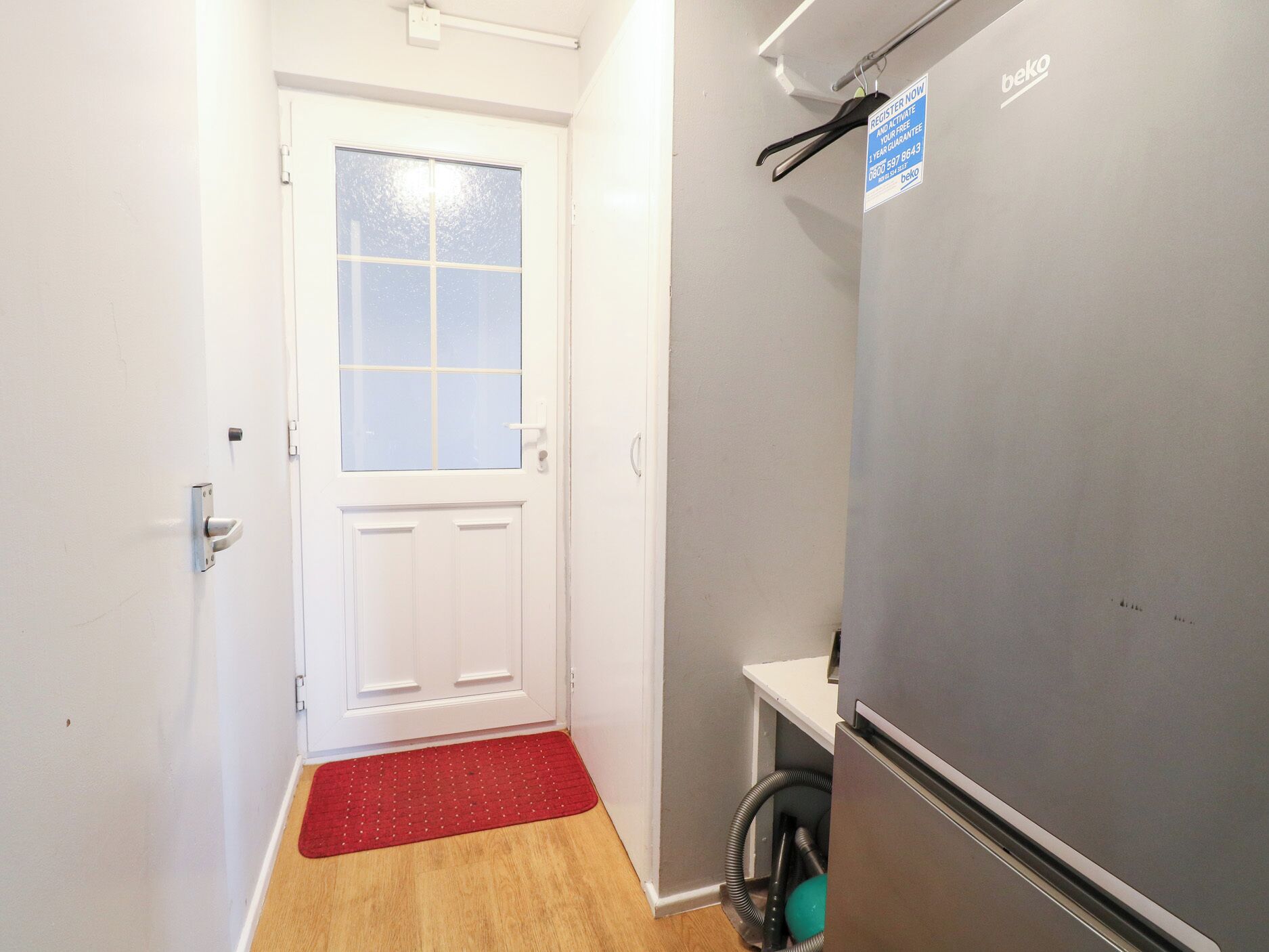A narrow hallway with a white door, a red doormat, a grey refrigerator, and a sign on the refrigerator.