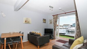 A living room with a wooden table, black sofa, and a TV. There are two paintings on the wall and a view of boats through the window.