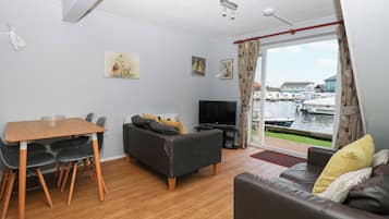 Ein Wohnzimmer mit Holztisch, schwarzem Sofa und Fernseher. Zwei Gemälde hängen an der Wand und durch das Fenster ist ein Blick auf Boote zu sehen.