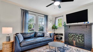 A modern living room with a leather sofa, a round coffee table, a fireplace with a tree design, and a ceiling fan. - Pet Friendly Cute Cottage - Walk to PC/Downtown (Clinton)