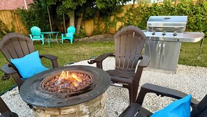 Un patio con una chimenea de piedra, dos sillas Adirondack con cojines azules y una parrilla.