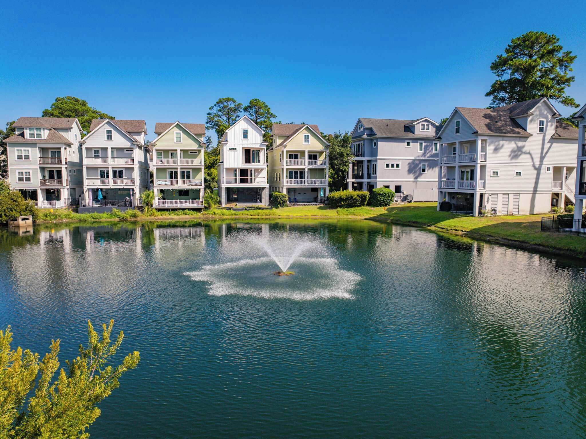 A row of multi-story residential buildings with balconies, a pond in the foreground, and a clear blue sky.