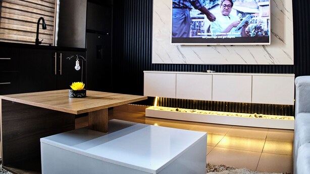 A modern living room with a flat-screen TV mounted on the wall, a wooden coffee table, a white sofa, and a kitchen area with dark cabinetry. - Blays Hideout - Cozy Apartments (Accra)