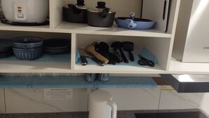 Eine Küche mit einem weißen Kühlschrank, einem weißen Slow Cooker, einer blauen Bratpfanne und verschiedenen Küchenutensilien in Regalen.