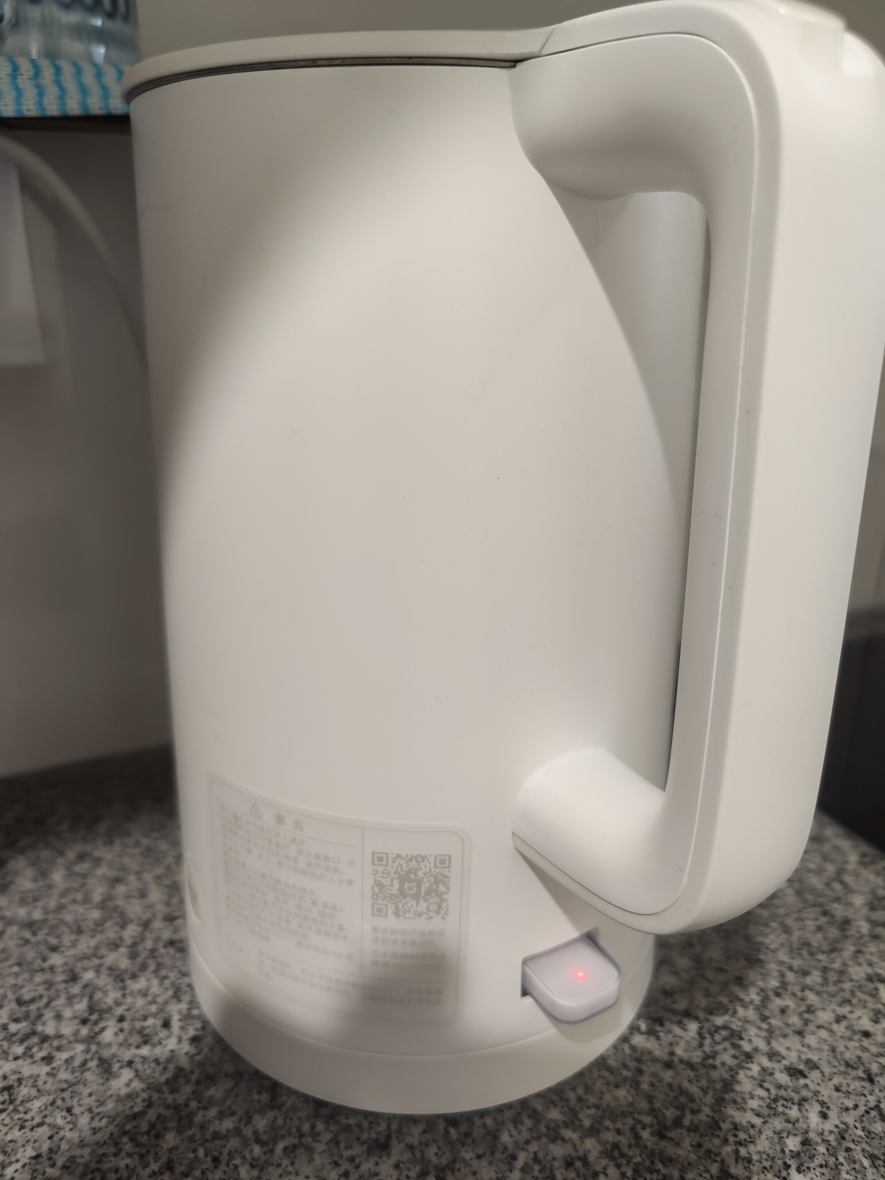 Una tetera eléctrica blanca con un código QR y un botón de encendido sobre una encimera de granito.