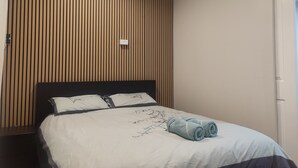 Ein ordentlich bezogenes Bett mit weiß-blauer Tagesdecke, einem dunklen Kopfteil und einer Wand mit vertikalen Holzhakobslatten.