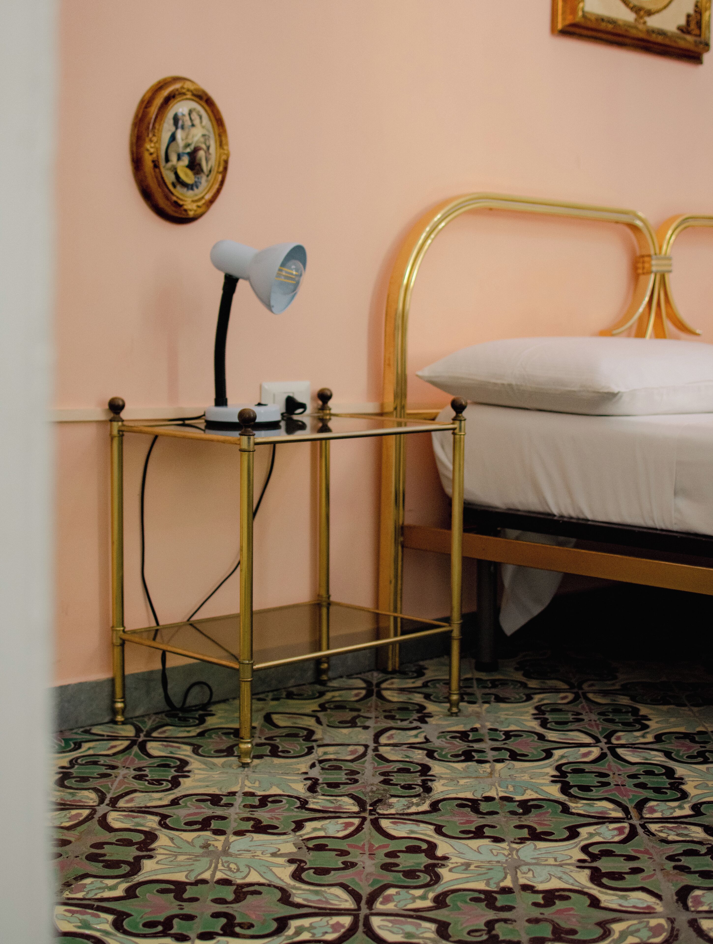A room with a bed, a bedside table with a lamp, a framed picture on the wall, and patterned floor tiles.