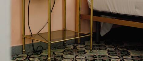 A room with a bed, a bedside table with a lamp, a framed picture on the wall, and patterned floor tiles.