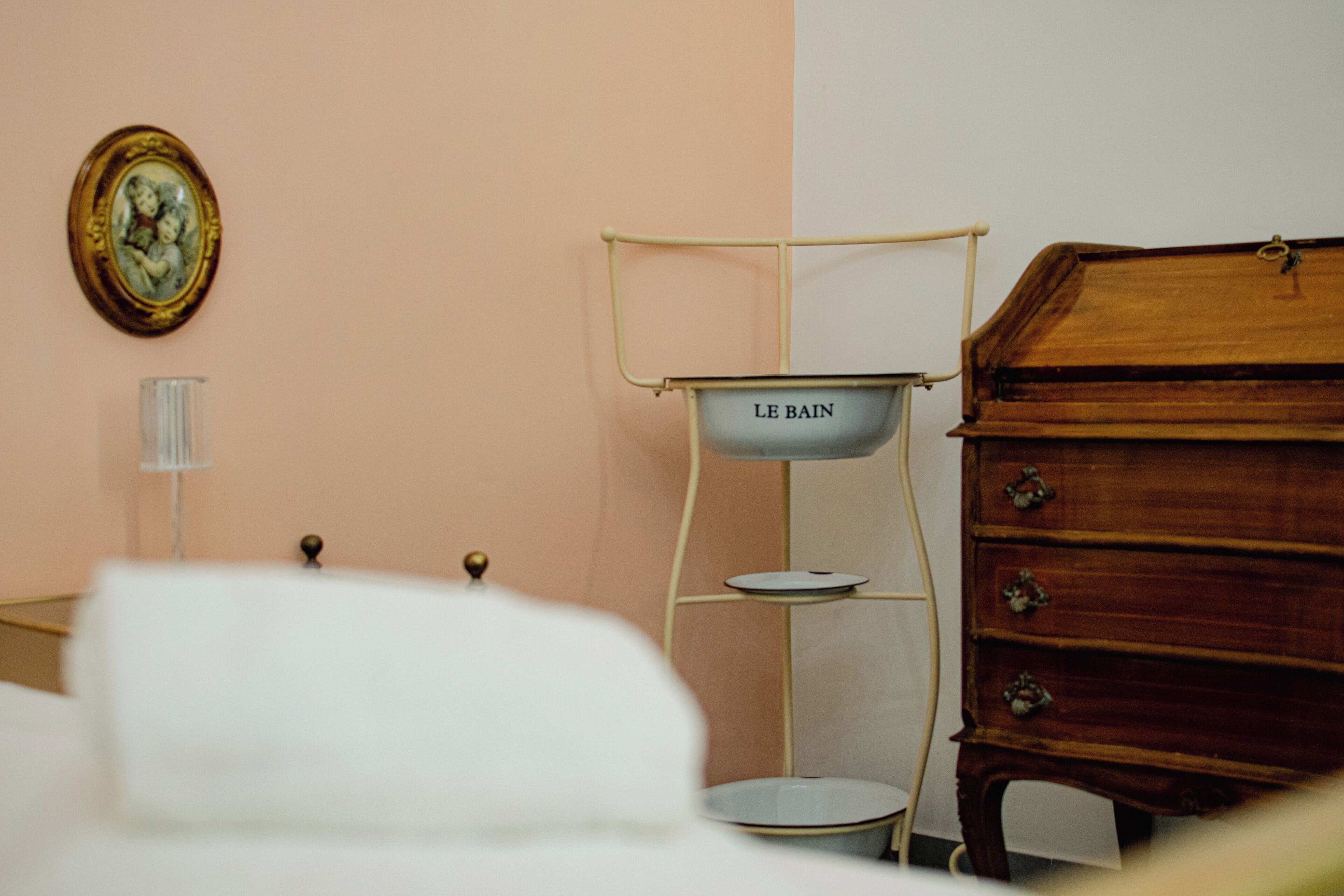 A bedroom with a bed, a dresser, a bedside table, a lamp, and a wall-mounted shelf with a basin.